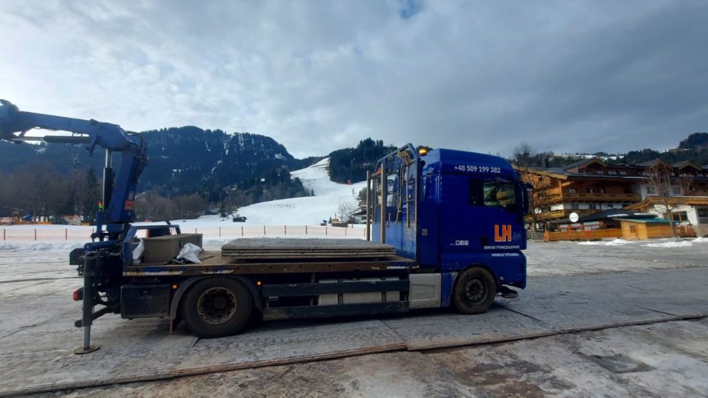 Niebieska ciężarówka na tle górskiego stoku narciarskiego, obok drewniane palety. Zimowa sceneria.
