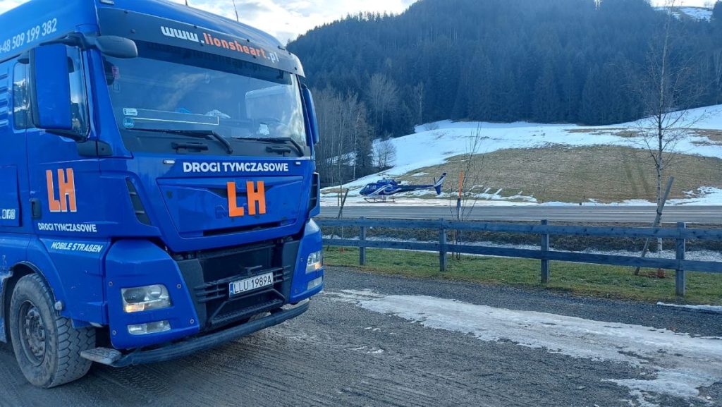 Niebieska ciężarówka z napisem LH na drodze, w tle helikopter i ośnieżone tereny górskie.