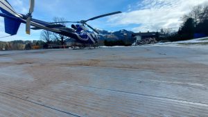 Helikopter na płycie lądowiska z widokiem na góry i niebo z chmurami. W tle widoczny dom i zieleń.