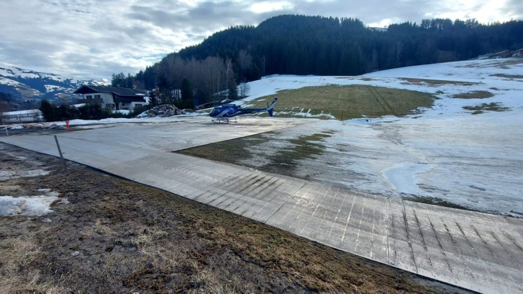 Lądowisko dla helikopterów na tle górskiego krajobrazu, z resztkami śniegu i zieloną trawą.