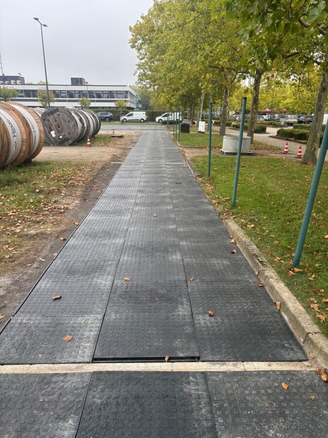 Fragment chodnika z czarnymi płytami, w tle widoczne walce i pojazdy, otoczony zielenią drzew.