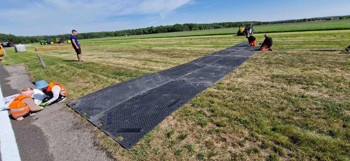 Pracownicy układają czarne panele na trawie, w tle maszyny rolnicze i zielone pola.
