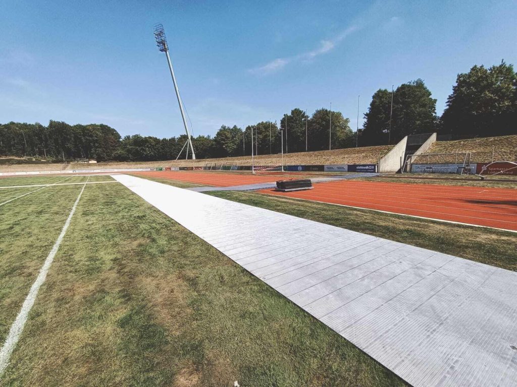 Widok na stadion z bieżnią, torami oraz prostą, białą nawierzchnią na boisku, w tle drzewa i słup oświetleniowy.