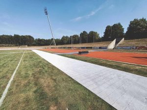 Widok na stadion z bieżnią, torami oraz prostą, białą nawierzchnią na boisku, w tle drzewa i słup oświetleniowy.