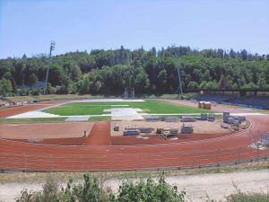 Boisko sportowe z bieżnią, w trakcie prac budowlanych, otoczone lasem i widocznymi materiałami budowlanymi.