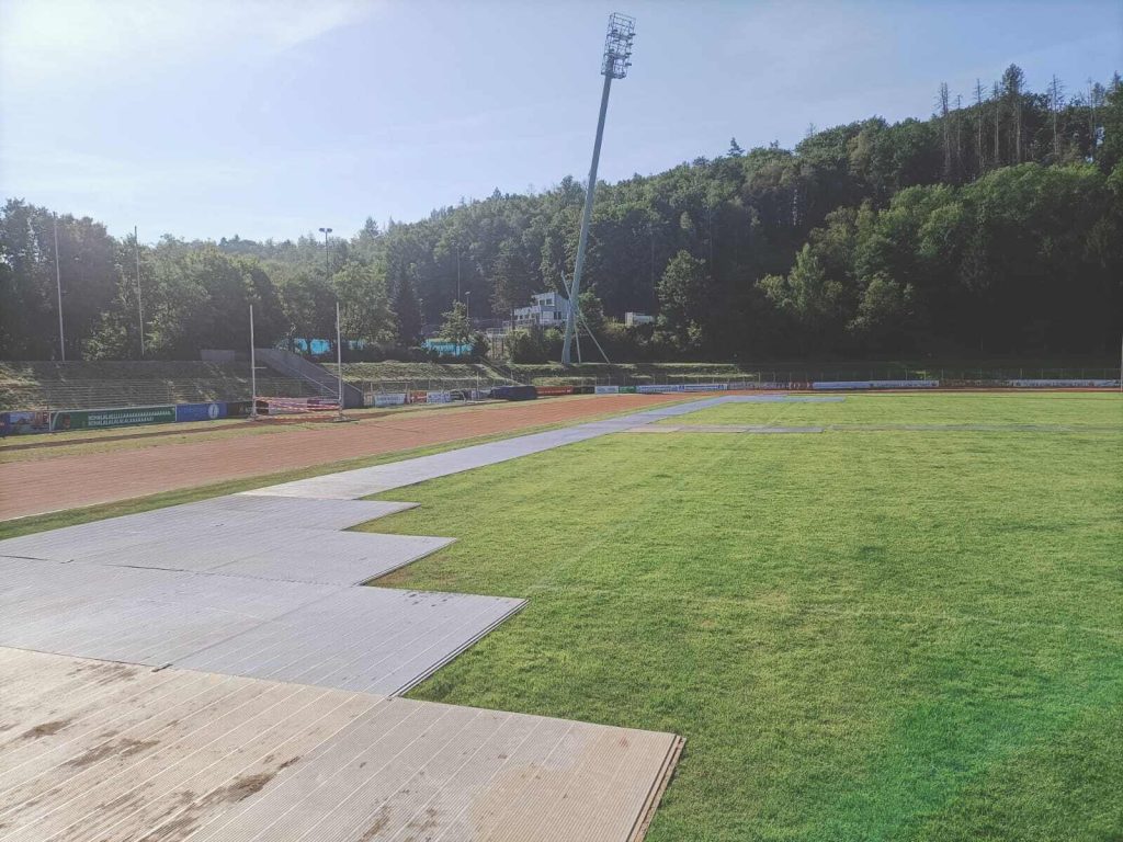 Widok na stadion z bieżnią, zieloną murawą oraz trybunami otoczonymi drzewami. W tle widać oświetlenie stadionowe.