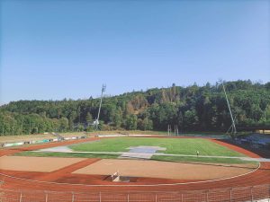 Pusty stadion lekkoatletyczny z bieżnią, boiskami i otaczającą go zielenią drzew. Na niebie bezchmurne słońce.