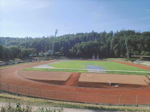 Widok na stadion sportowy z bieżnią, boiskiem trawiastym oraz otaczającą zieleń drzew.