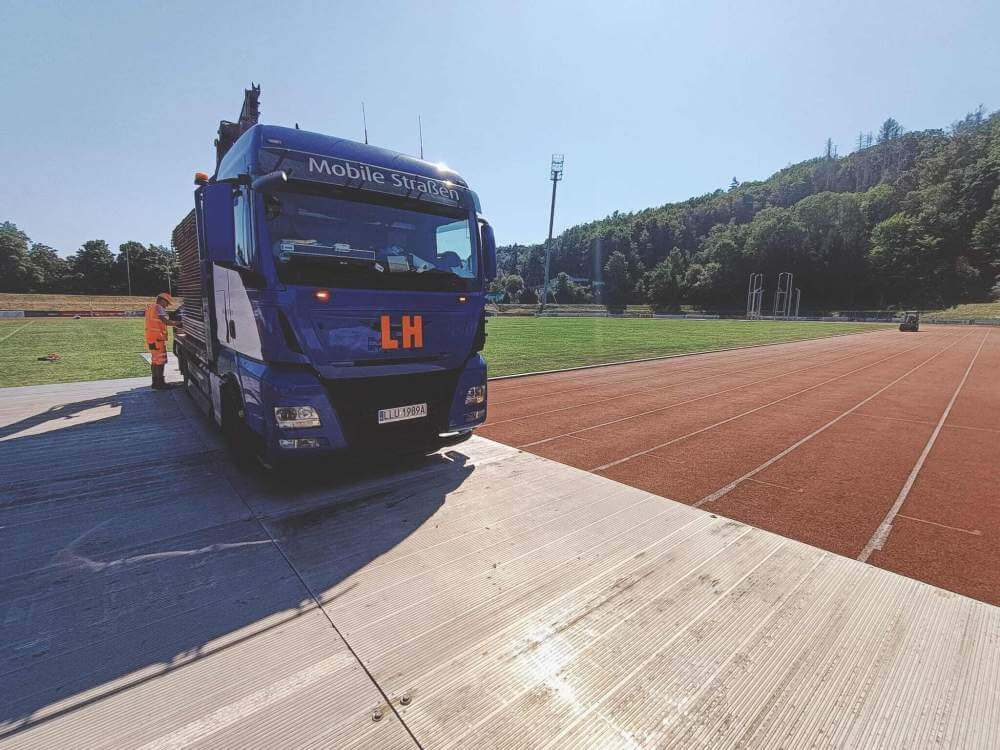 Niebieska ciężarówka na stadionie lekkoatletycznym, obok bieżni, w tle zieleń drzew. Słońce świeci jasno.