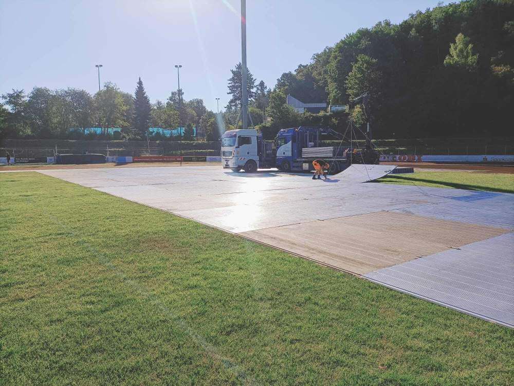 Na zdjęciu widać ciężarówkę oraz pracownika układającego panele na trawie na boisku. W tle rosną drzewa.
