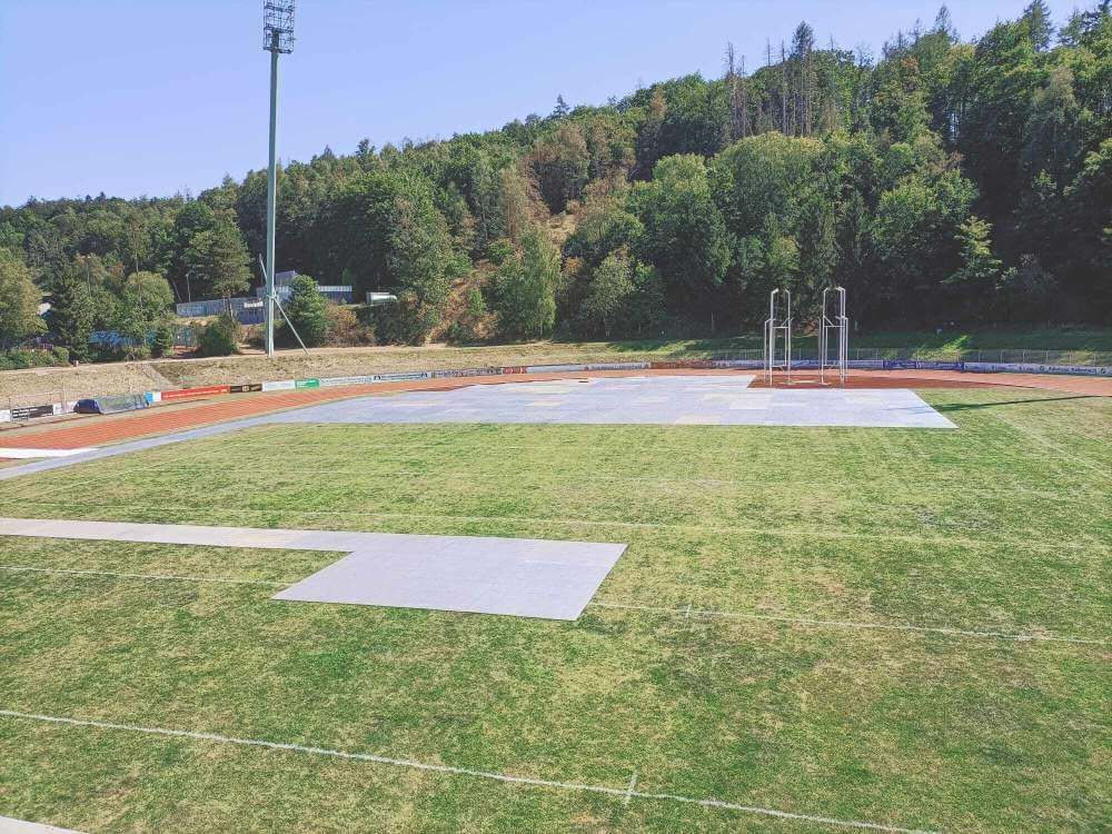 Pusty stadion lekkoatletyczny z zieloną murawą, widocznymi torami biegowymi i bieżnią, otoczony lasem.