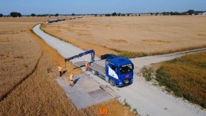 Ciężarówka na drodze w polu, dwóch pracowników w kamizelkach odblaskowych przy wózku. W tle zboże i niebo.