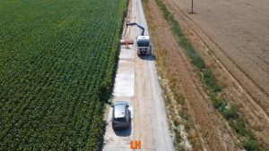 Na obrazie widać drogę wiejską z samochodem po lewej stronie, a po prawej stronie pola kukurydzy oraz ciężarówkę z dźwigiem.