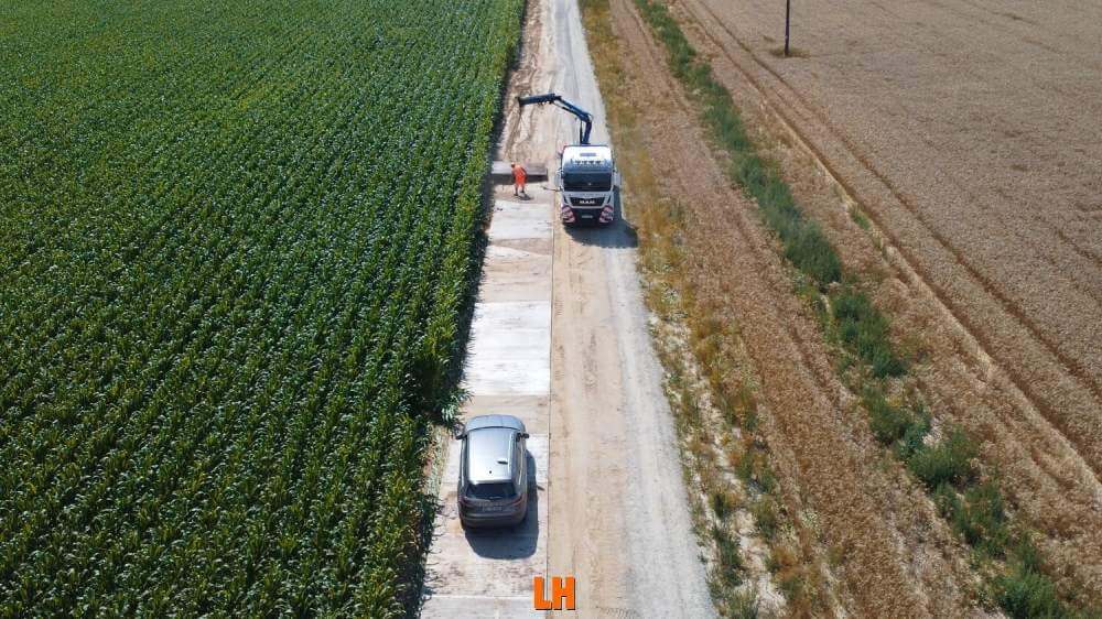 Na obrazie widać drogę wiejską z samochodem po lewej stronie, a po prawej stronie pola kukurydzy oraz ciężarówkę z dźwigiem.