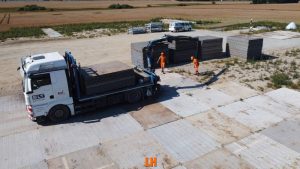 Na obrazku widać ciężarówkę z załadunkiem na placu budowy, dwóch pracowników w pomarańczowych strojach oraz sterty płyt.