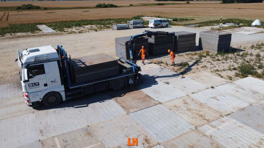 Na obrazku widać ciężarówkę z załadunkiem na placu budowy, dwóch pracowników w pomarańczowych strojach oraz sterty płyt.