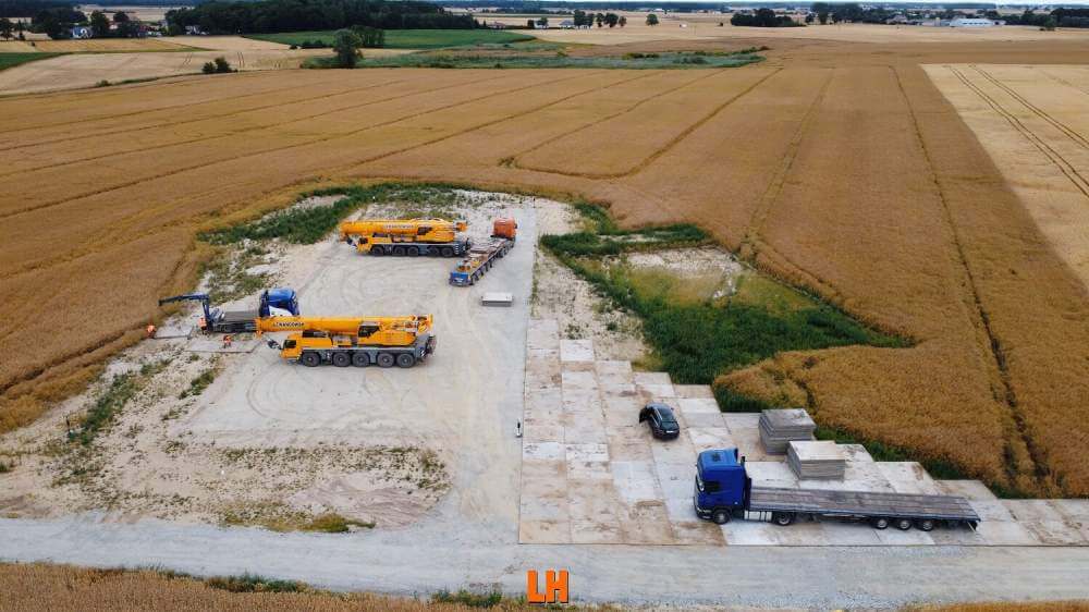 Na zdjęciu widać plac budowy z ciężkim sprzętem, przyczepą i materiałami budowlanymi na tle złotych pól.