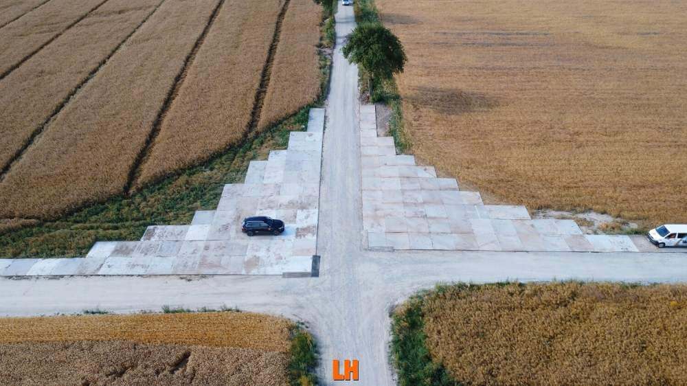 Widok z góry na skrzyżowanie dróg wyłożonych betonem w otoczeniu pól zbożowych. Widać dwa samochody stojące na drodze.