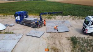 Ciężarówka z dźwigiem podnosi panele na placu budowy w otoczeniu pól. Pracownicy w kamizelkach odblaskowych.