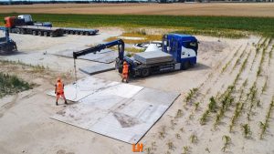 Na obrazie widoczny jest niebieski ciężarówka z podnośnikiem oraz dwie osoby w pomarańczowych kamizelkach, układające płyty n