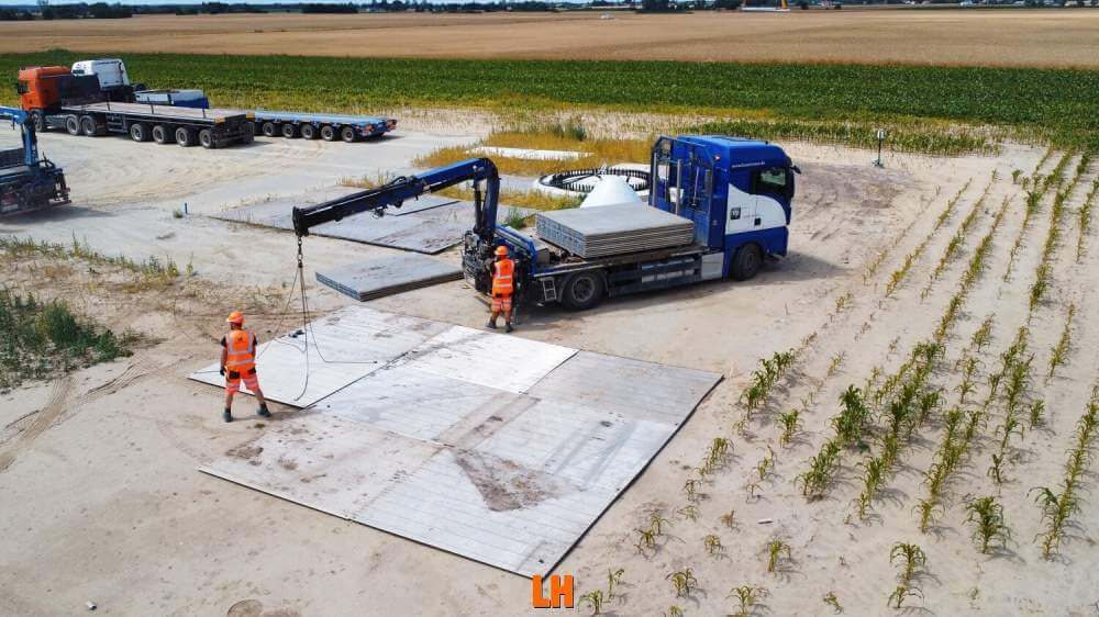 Na obrazie widoczny jest niebieski ciężarówka z podnośnikiem oraz dwie osoby w pomarańczowych kamizelkach, układające płyty n