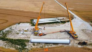 Obraz przedstawia dwa żurawie montażowe przy wietraku oraz leżące na ziemi łopaty wirnika na polu.