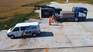 Grupa pracowników na placu budowy, samochód dostawczy i ciężarówka z kontenerem w tle.