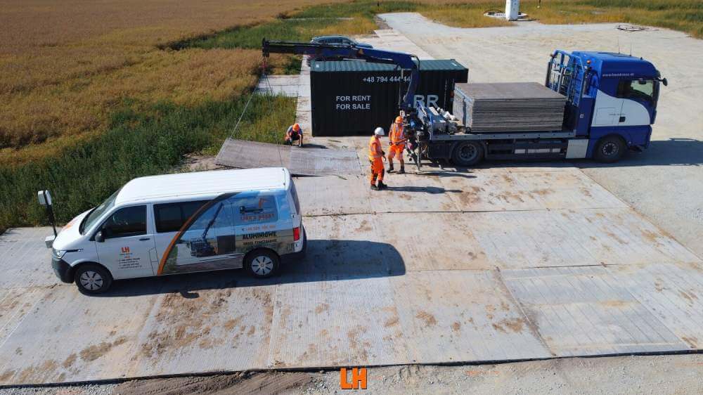 Grupa pracowników na placu budowy, samochód dostawczy i ciężarówka z kontenerem w tle.