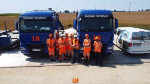 Grupa pracowników w pomarańczowych strojach roboczych stoi przed dwoma niebieskimi ciężarówkami na tle pola.