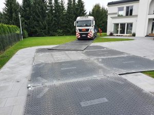 Tir na podjeździe, na którym znajduje się rozłożona perforowana płyta, prowadząca do budynku. W tle drzewa.