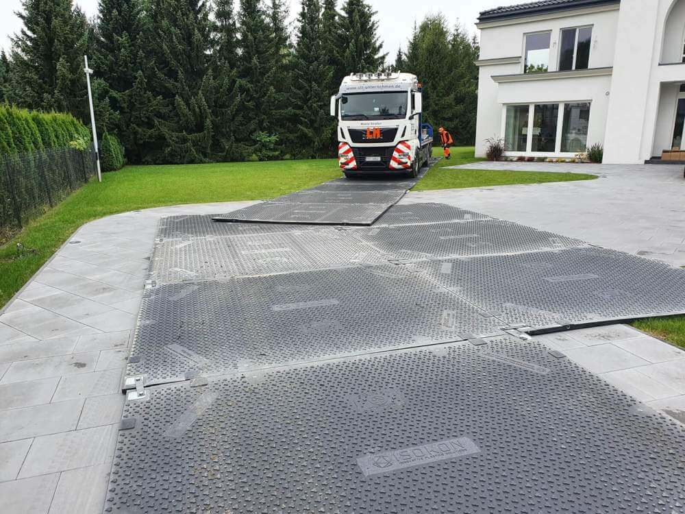 Tir na podjeździe, na którym znajduje się rozłożona perforowana płyta, prowadząca do budynku. W tle drzewa.