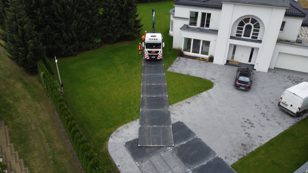 Czerwony pojazd na podjeździe nowoczesnego domu, obok parked biały van, trawa w tle, drzewa po lewej.