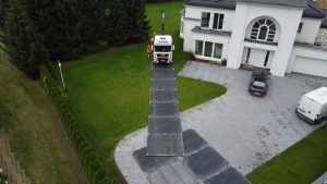 Czerwony pojazd na podjeździe nowoczesnego domu, obok parked biały van, trawa w tle, drzewa po lewej.