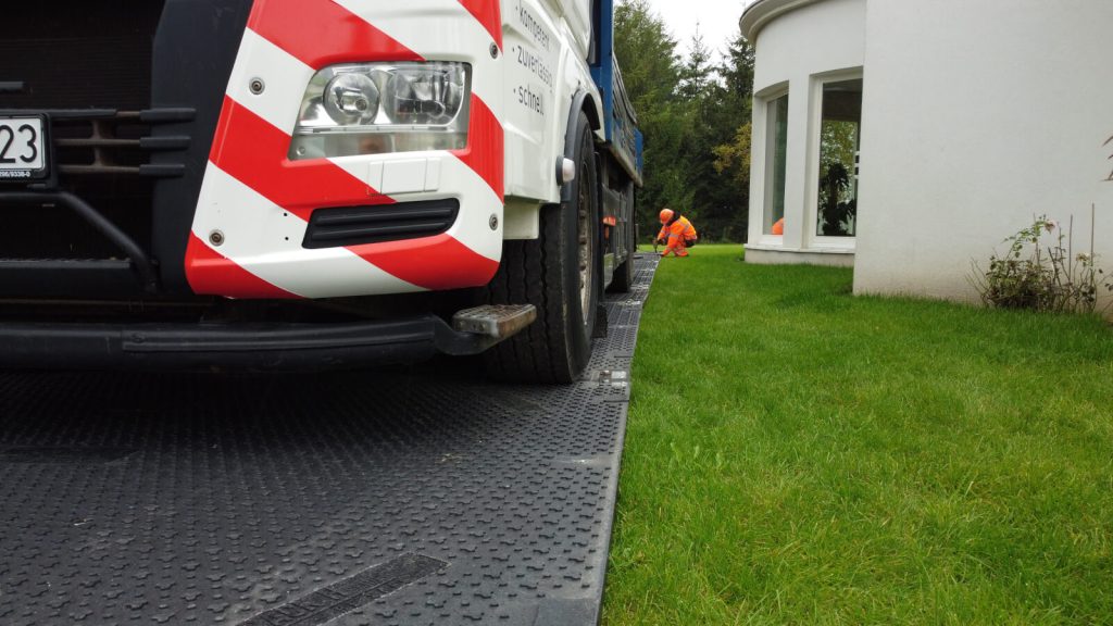 Ciężarówka na drodze z pasami ostrzegawczymi, obok pracownik w pomarańczowej odzieży, w tle trawniki i budynek.