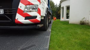 Ciężarówka na drodze z pasami ostrzegawczymi, obok pracownik w pomarańczowej odzieży, w tle trawniki i budynek.