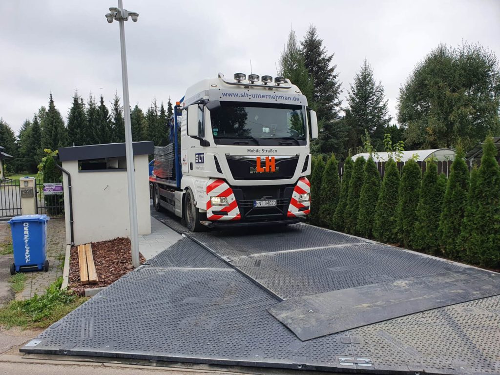 Ciężarówka stoi na platformie wjazdowej przed budynkiem, otoczona drzewami i koszami na odpady.