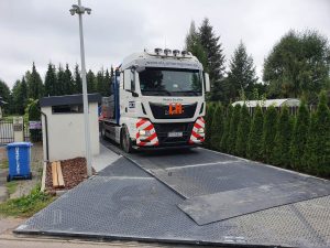 Ciężarówka stoi na platformie wjazdowej przed budynkiem, otoczona drzewami i koszami na odpady.