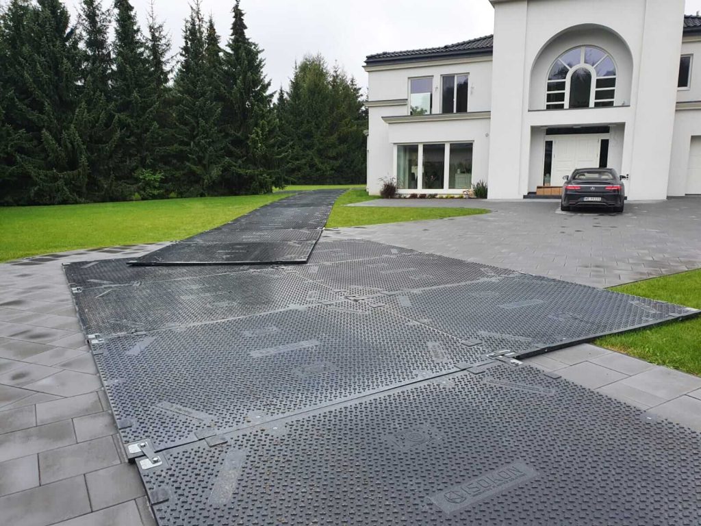 Wjazd do nowoczesnego domu z czarną kostką brukową, pokryty matami ochronnymi, obok parkowany samochód.