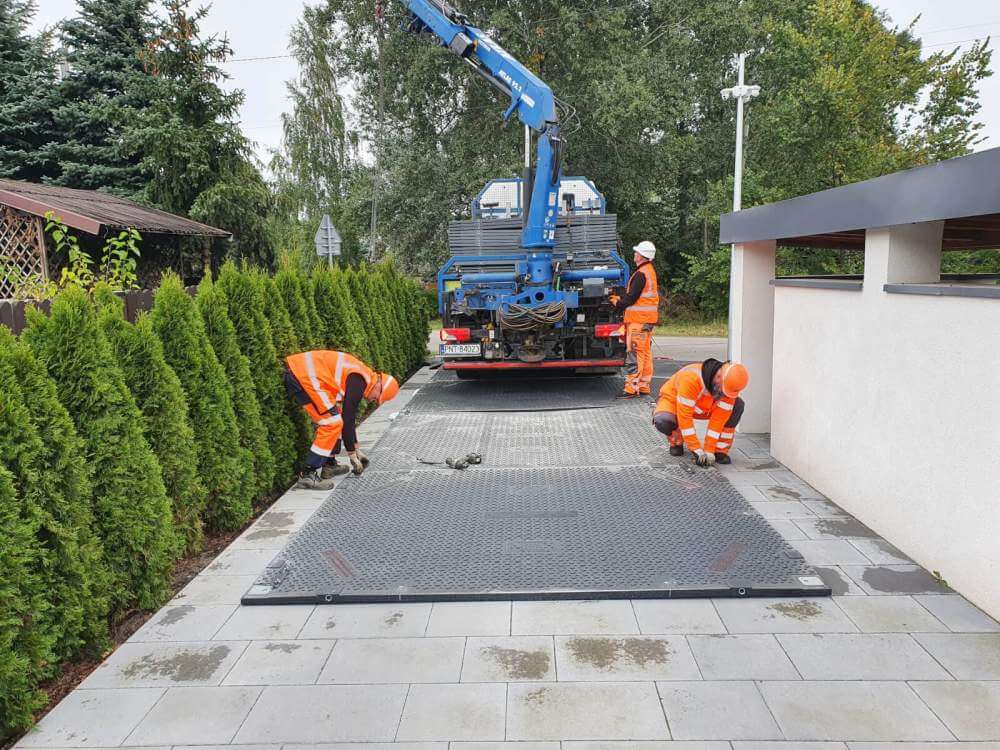 Pracownicy w odblaskowych ubraniach montują elementy na placu budowy, obok podniesiony dźwig.