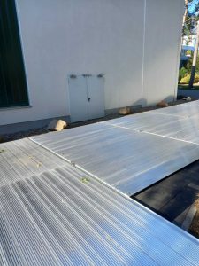 Szeroka, metalowa powierzchnia paneli słonecznych na podłożu z ciemnego materiału obok budynku.