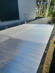 Szeroka, metalowa nawierzchnia obok budynku, otoczona trawnikiem i drzewami.