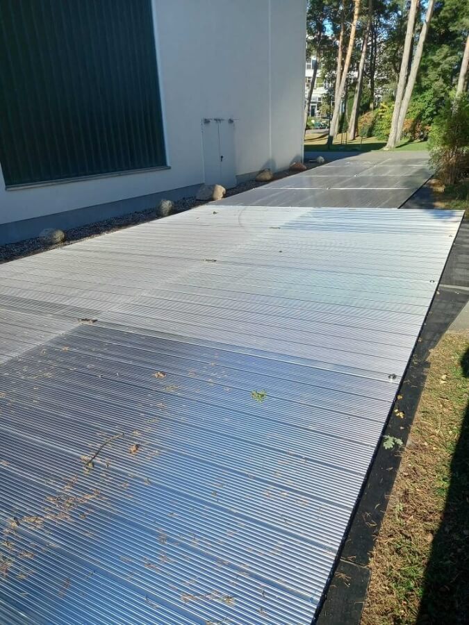 Szeroka, metalowa nawierzchnia obok budynku, otoczona trawnikiem i drzewami.