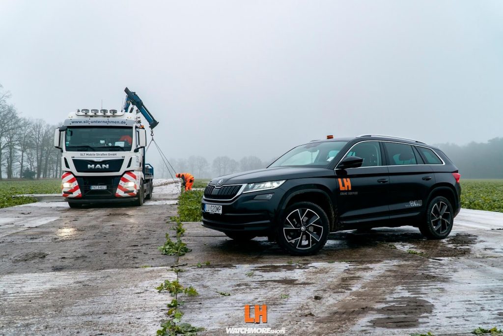 Czarny SUV stojący na drodze obok ciężarówki MAN, w tle widoczna mgła i pracownik przy maszynie.