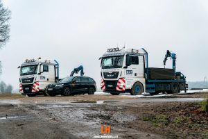 Dwóch ciężarówek z podnośnikami obok samochodu osobowego na drodze w deszczowy dzień. W tle widoczna szarość nieba.