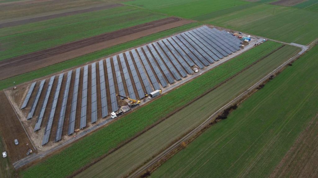 Widok z lotu ptaka na farmę słoneczną z panelami słonecznymi na zielonym polu. W tle widać maszyny budowlane.