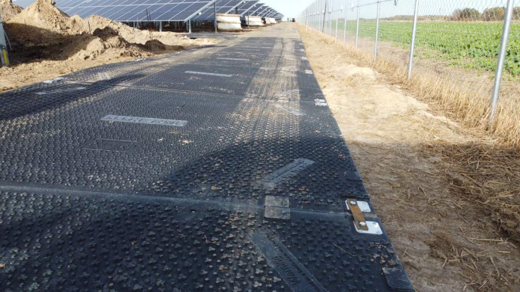 Droga wyłożona panelami w farmie fotowoltaicznej, otoczona polem i ogrodzeniem. W tle widać rzędy paneli słonecznych.