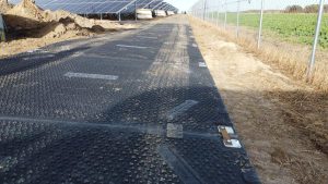 Droga wyłożona panelami w farmie fotowoltaicznej, otoczona polem i ogrodzeniem. W tle widać rzędy paneli słonecznych.