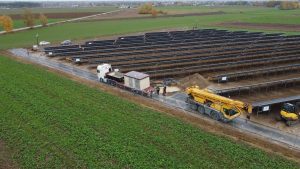 Pole z panelami słonecznymi, z pojazdami i pracownikami przy budowie lub instalacji. W tle zielone tereny.