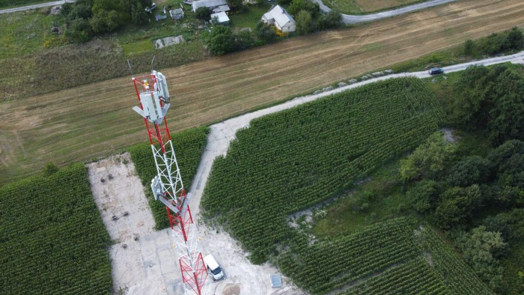 Widok na wieżę telekomunikacyjną otoczoną polami i drzewami, w tle widać kilka budynków oraz drogę.