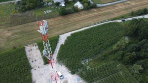 Widok na wieżę telekomunikacyjną otoczoną polami i drzewami, w tle widać kilka budynków oraz drogę.